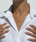 A close-up lifestyle shot of a model wearing a delicate gold necklace with a small clear crystal pendant, styled with a white button-down shirt.